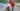 Foto colorida de mulher de pele escura e cabelos grisalhos vestindo camisa de manga longa vermelha e saia estampada segurando um balde com mariscos de forma inclinada para que caiam dentro de uma panela sobre um fogão à lenha improvisado