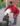 Foto colorida de mulher de pele escura e cabelos grisalhos vestindo camisa de manga longa vermelha e saia estampada segurando um balde com mariscos de forma inclinada para que caiam dentro de uma panela sobre um fogão à lenha improvisado