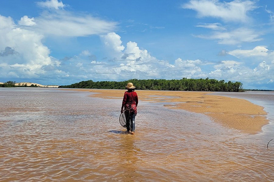Foto colorida de rio na maré baixa com mulher de costas e em pé usando chapéu de palha e blusa vermelha segurando uma rede de pesca de frente para vegetação de mangue e areial