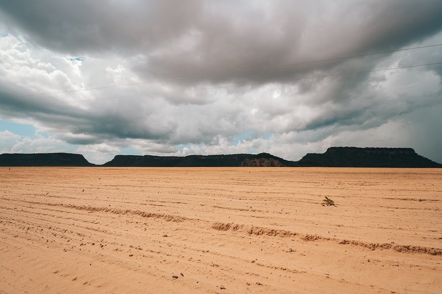 Foto colorida de extensão área de solo cor de barro sendo preparada para receber plantação agrícola tendo ao fundo um morro que se espalha no horizonte sob um céu coberto de nuvens cinzas