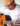 Fotografia de um homem pardo, usando óculos de graus, boina, de cor ocre, blusa branca misturada a outros tons claros. O homem segura um violão de seis cordas, enquanto toca o instrumento. Ao fundo há uma parede verde clara