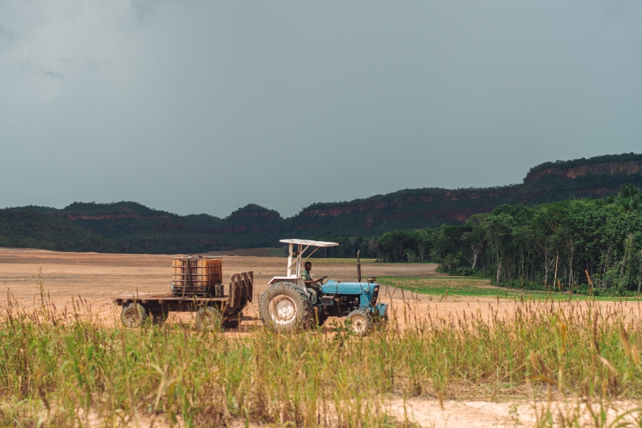 Foto colorida de trator em deslocamento em área de plantio tendo ao fundo vegetação nativa de Cerrado e um platô coberto de vegetação