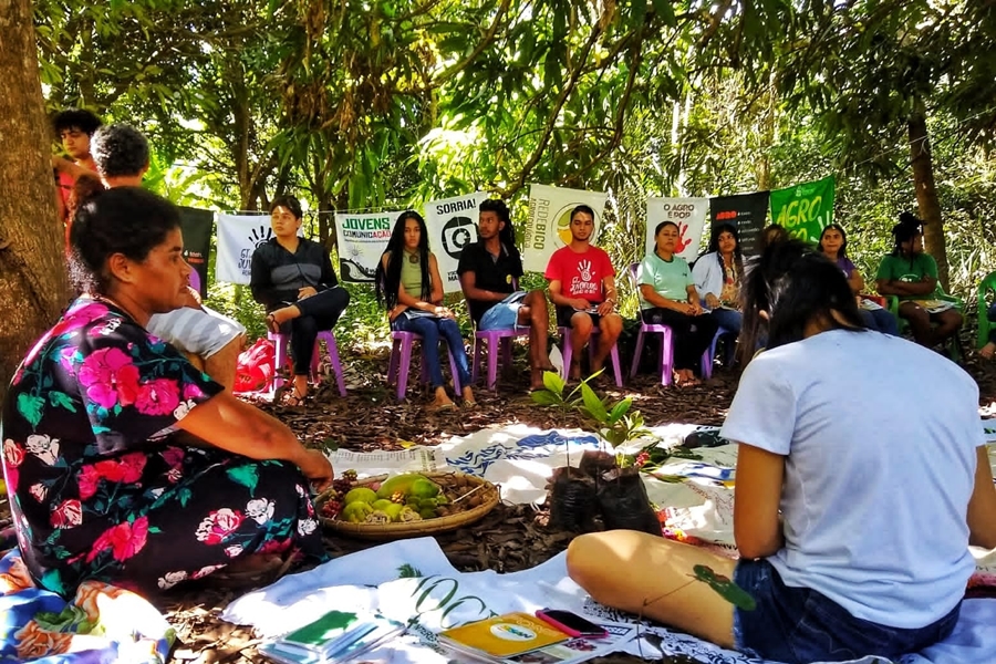 Foto colorida de grupo de pessoas sentadas em círculo tendo em volta cartazes pendurados e ao centro mudas de plantas e mais cartazes dispostos no chão.