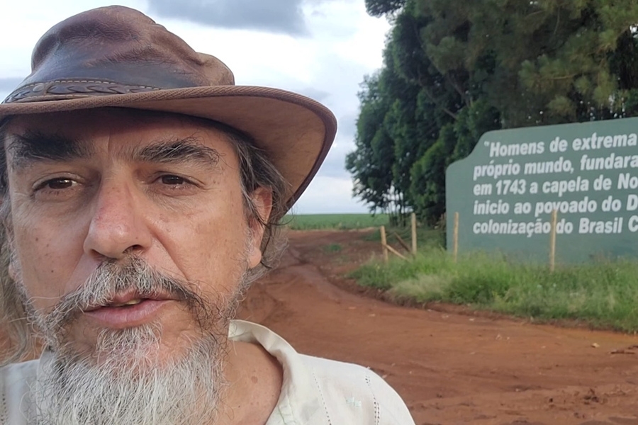 Foto colorida de homem branco de bigode e barba grisalhos usando chapéu e tendo atrás de si uma estrada de barro vermelho, árvores e plantação ao fundo