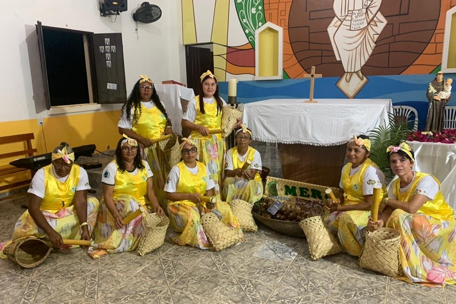 Foto colorida de mulheres vestidas com saias e blusas amarelas segurando quebradores de coco babaçu sentadas em frente a um altar de igreja católica com crucifixo e imagem de Santo Antônio