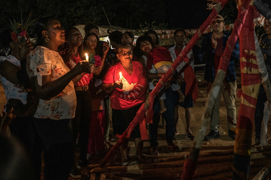 Foto feita em ambiente aberto, à noite, de várias pessoas ao redor de uma estrutura feita com trancos finos cobertos com fitas brancas e vermelhas. Duas mulheres mais à frente seguram velas acesas. A mulher mais ao centro usa óculos e uma camiseta vermelha com o desenho da pomba branca que simboliza o divino espírito santo