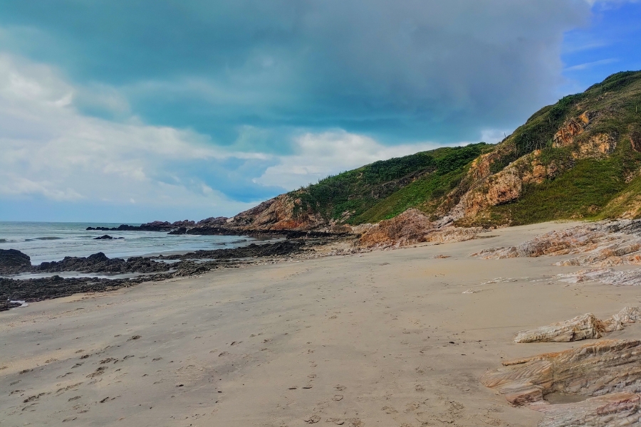 Foto de paisagem litorânea feita da faixa de areia. Do lado esquerdo, o mar com formações rochosas. À direita, falésias com vegetação