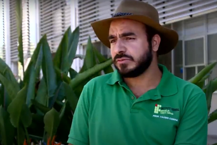 Foto colorida de homem pardo com barba e bigode pretos usando chapéu marrom e camisa polo verde onde se lê na altura do peito Instituto Federal de Brasília Prof. Tássio Cunha