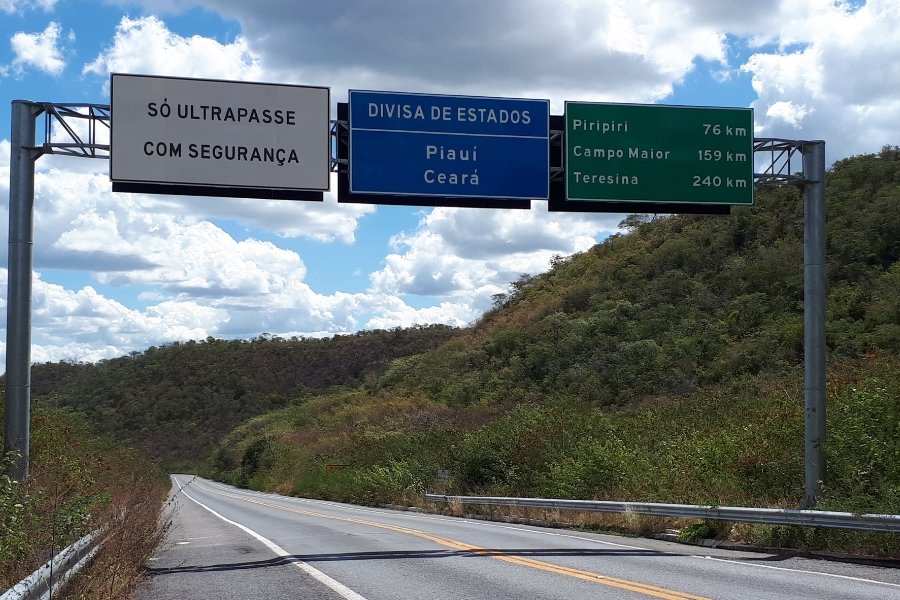 Foto de estrada asfaltada ladeada por elevações cobertas de vegetação. Há uma estrutura metálica sobre a rodovia com três placas. Na primeira, branca com letras pretas, lê-se: 'SÓ ULTRAPASSE COM SEGURANÇA'; na segunda, azul com letras brancas, 'DIVISA DE ESTADOS Piauí Ceará; na terceira, verde com letras brancas, Piripiri 76km Campo Maior 159km Teresina 240km'