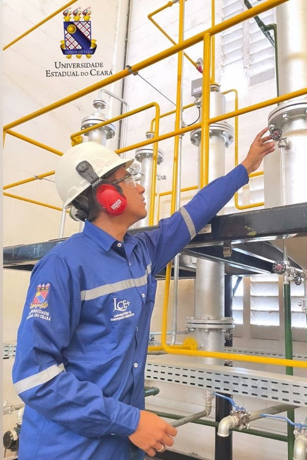 Foto de homem trajando uniforme azul, capacete branco e fones de ouvido vermelhos e manipulando válvula em grande tanque branco com canos externos amarelos. Na parede ao fundo, brasão e UNISERSIDADE ESTADUAL DO CEARÁ