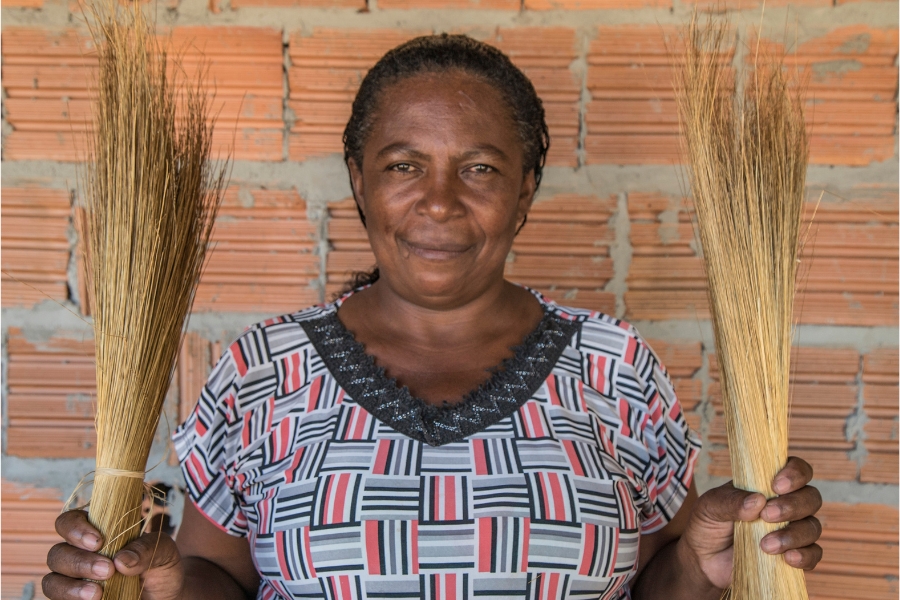 Mulher negra sorridente pousa para foto à frente de uma parede de tijolos aparentes segurando dois feixes de capim dourado. Ela tem os cabelos escuros amarrados em um coque e usa blusa estampada de quadrados e listras pretas, azuis, brancas e rosa