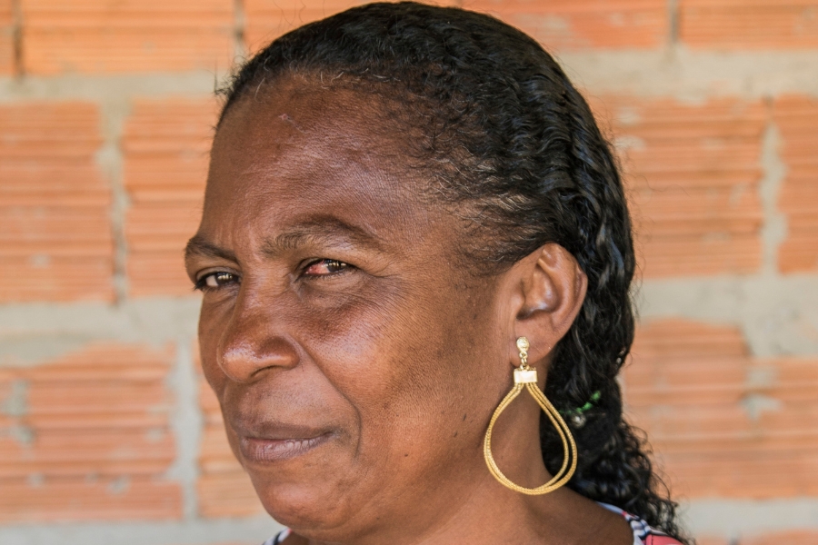 Foto colorida de mulher negra de cabelos escuros e amarrados em um rabo de cavalo de perfil olhando de lado para a câmera e exibindo um brinco artesanal feito de capim dourado