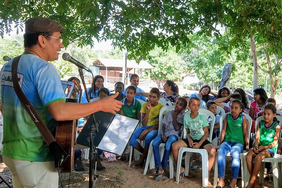 Foto colorida de homem tocando violão e cantando diante de plateia composta por crianças sentadas em cadeiras plásticas