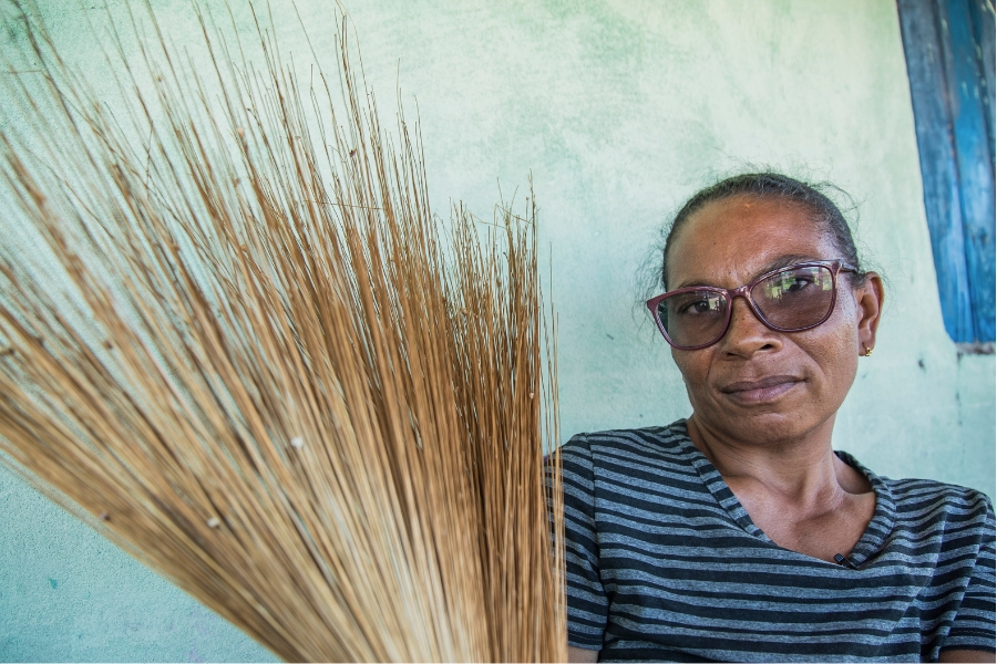 Foto colorida de mulher negra de óculos de grau e cabelos escuros amarrados em um coque olhando de frente para a câmera e segurando com a mão direita um feixe de capim dourado