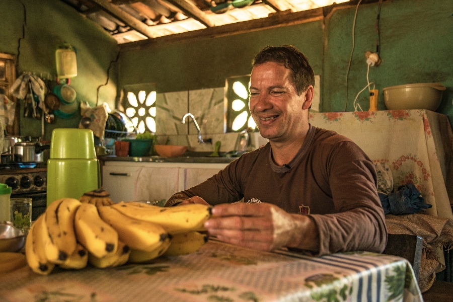 Foto colorida de homem pardo usando blusa de mangas compridas marrom sentado à mesa em uma cozinha sorrindo enquanto mostra uma palma de banana madura