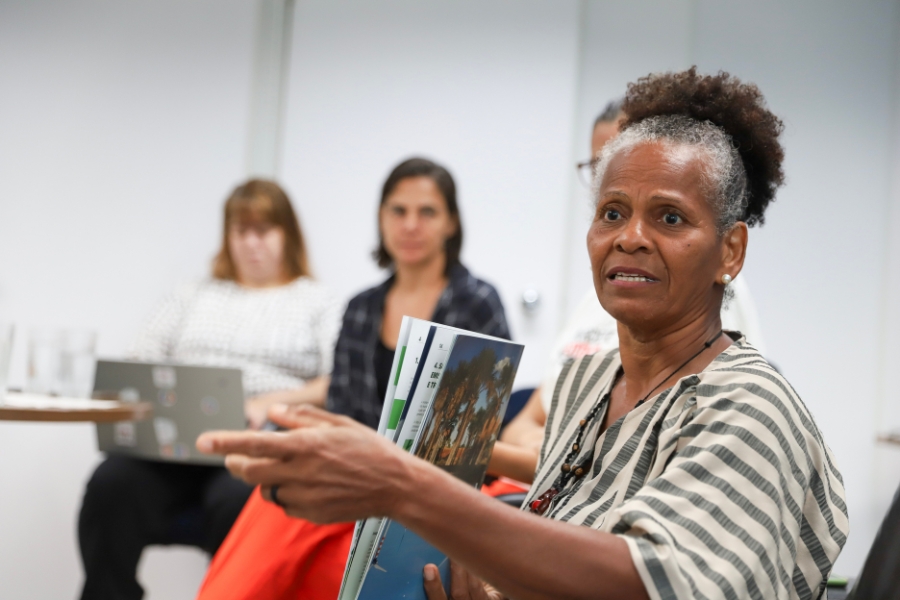 Foto colorida de mulher negra usando blusa de listras brancas e verdes e cabelo amarrado no alto da cabeça. Ela segura um livro e está sentada ao lado de outras duas mulheres que aparecem desfocadas na imagem