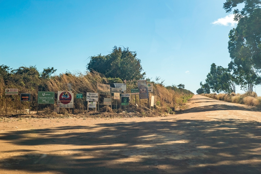 Foto colorida de estrada de barro ladeada por vegetação esparsa com dezenas de placas indicativas de fazendas localizadas na região