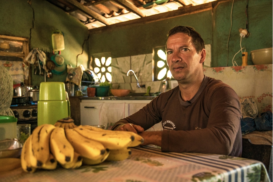 Foto colorida de homem pardo de camisa marrom sentado à mesa de uma cozinha olhando para a câmera e tendo à frente uma palma de banana madura e uma garrafa de café de plástico verde claro