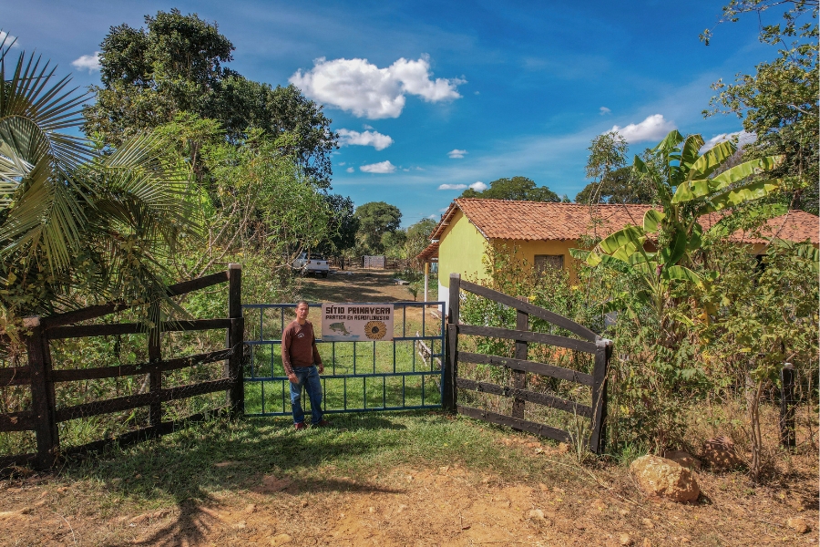 Foto colorida de homem pardo de camisa marrom e calça jeans em frente a porteira com placa onde se lê Sitio Primavera tendo atrás uma casa amarela coberta de telhas de argila e cercada de árvores e jardins