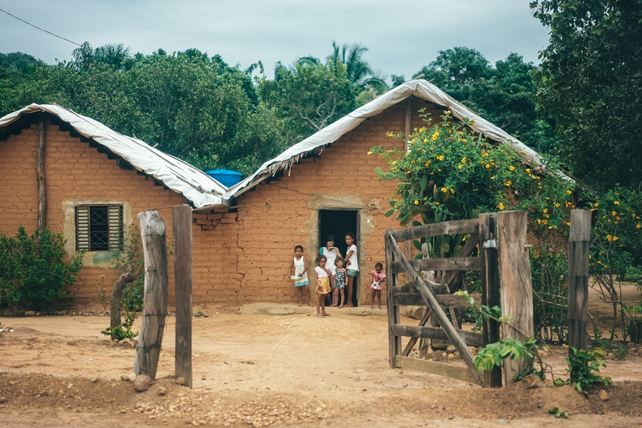 Foto colorida de família de mulheres adultas e crianças pousando na frente de uma casa de paredes de barro e com porteira aberta