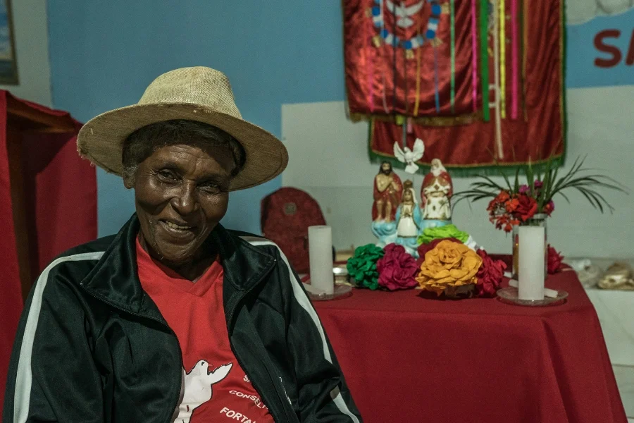 Foto colorida de mulher negra e sorridente de chapéu de palha, blusa vermelha e casaco preto sentada à frente de um altas caseiro com imagens de santos católicos