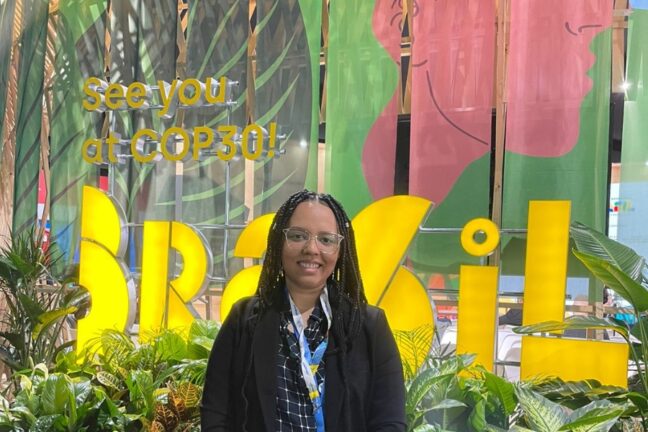 A imagem mostra uma mulher sorridente posando em frente a um estande com a mensagem em inglês "See you at COP30!" (Vejo você na COP30!) e a palavra "Brazil" destacada em letras grandes e amarelas. O cenário é decorado com muitas plantas, representando um ambiente tropical ou sustentável, enquanto ao fundo há elementos visuais com ilustrações estilizadas e coloridas. A pessoa usa roupas formais e um crachá, indicando participação oficial em um evento, possivelmente relacionado à COP29, com referência à próxima conferência (COP30). O estande promove o Brasil como próximo anfitrião do evento.