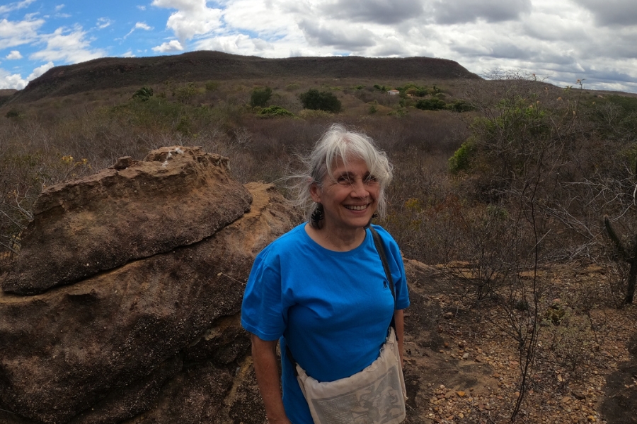 A imagem mostra uma mulher de cabelos grisalhos em pé ao ar livre em uma paisagem seca e rochosa com vegetação esparsa. A pessoa está vestindo uma camisa azul e carrega uma bolsa a tiracolo. Ao fundo, há colinas e um céu parcialmente nublado. A cena parece estar em uma região semiárida ou árida, possivelmente durante a estação seca, devido à vegetação marrom e seca.