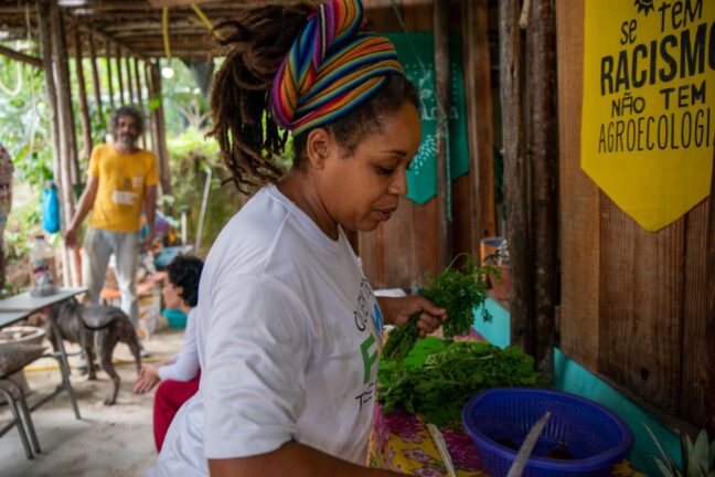 A imagem retrata uma mulher preparando ervas ou vegetais frescos em um ambiente rústico, que parece ser uma cozinha ou área de preparo comunitária. Ela usa uma camiseta branca com uma estampa e está com um turbante colorido vibrante nos cabelos. Ao fundo, é possível ver outras pessoas interagindo, incluindo um homem com uma camiseta amarela e um cachorro, criando um clima descontraído. Na parede de madeira, há um cartaz amarelo com a frase: "Se tem racismo, não tem agroecologia", indicando um contexto de luta por justiça social e ambiental. A cena evoca um ambiente comunitário que valoriza práticas agroecológicas e inclusão social.