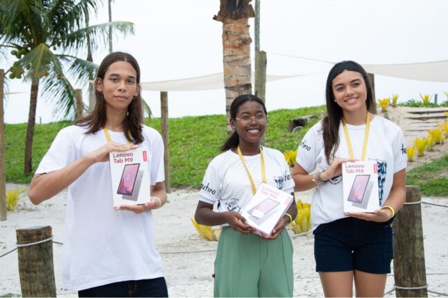 A imagem mostra três jovens sorridentes ao ar livre, segurando caixas de tablets Lenovo Tab M9. Eles estão vestidos com camisetas brancas personalizadas e cordões com credenciais pendurados no pescoço. O fundo apresenta um cenário de praia com coqueiro e grama verde.