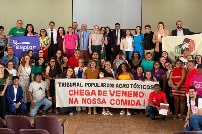 A imagem mostra um grande grupo de pessoas reunidas em uma sala, segurando faixas e cartazes. A faixa central diz: "TRIBUNAL POPULAR DOS AGROTÓXICOS CHEGA DE VENENO NA NOSSA COMIDA!" Há também outras duas faixas, uma à esquerda com o texto "esplar" e outra à direita com um logotipo e texto parcialmente oculto.