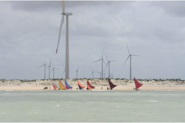 Foto colorida feita do mar mostra jangadas de pescadores atracando em praia tendo atrás cenário de dunas cobertas de turbinas eólicas