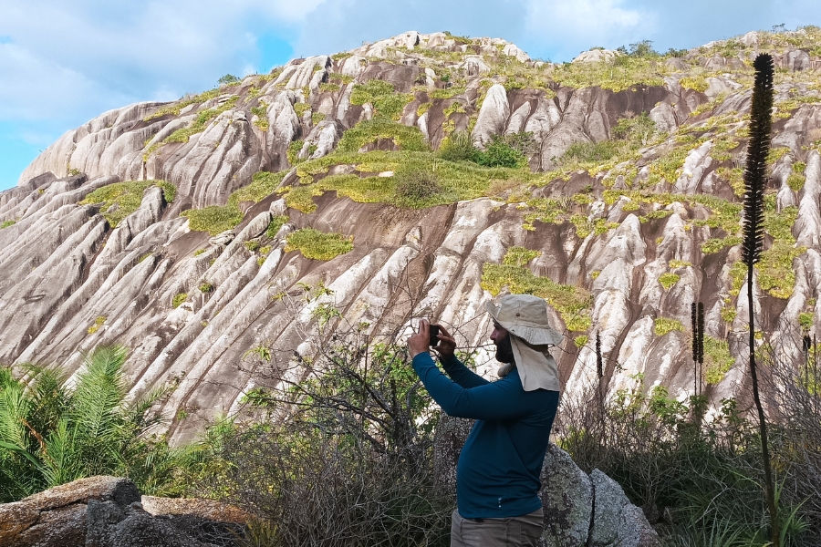 A imagem mostra uma paisagem natural impressionante com uma formação rochosa de grande porte ao fundo. A rocha possui um padrão único de sulcos e fendas verticais, com vegetação esparsa e verde crescendo entre as fendas, o que cria um contraste interessante com o tom acinzentado das pedras. Em primeiro plano, há um homem vestido com roupas apropriadas para trilha — uma camisa azul de manga comprida e um chapéu de proteção com abas largas para o sol. Ele está segurando um celular, aparentemente tirando uma foto ou filmando a paisagem à sua frente. Ao redor, há vegetação típica de ambientes semiáridos, com plantas secas, arbustos e um caule alto e escuro à direita da imagem, que pode ser de uma planta adaptada a ambientes secos. O céu está parcialmente nublado, com partes azuis visíveis entre as nuvens, o que sugere um clima ameno