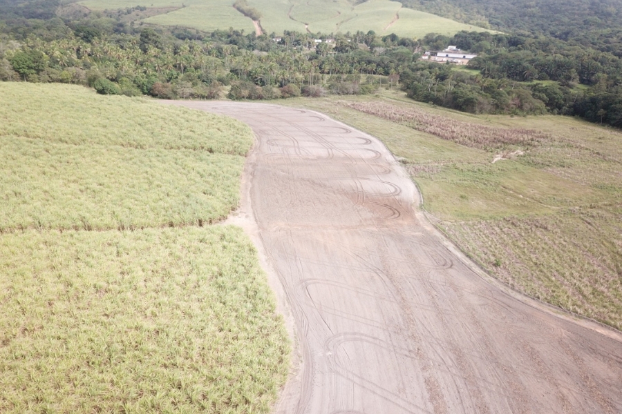 A imagem mostra uma área degradada com solo exposto e marcas de pneus, ao lado de plantações e vegetação nativa. Ao fundo, é possível ver uma comunidade rural cercada por morros verdes