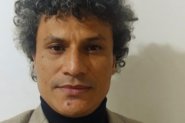 Foto close-up de um homem de meia-idade com cabelos cacheados escuros com alguns fios grisalhos. Ele tem um tom de pele moreno claro, olhos castanhos e lábios finos. Ele usa uma gola alta preta e um blazer bege claro. O fundo é uma parede branca lisa