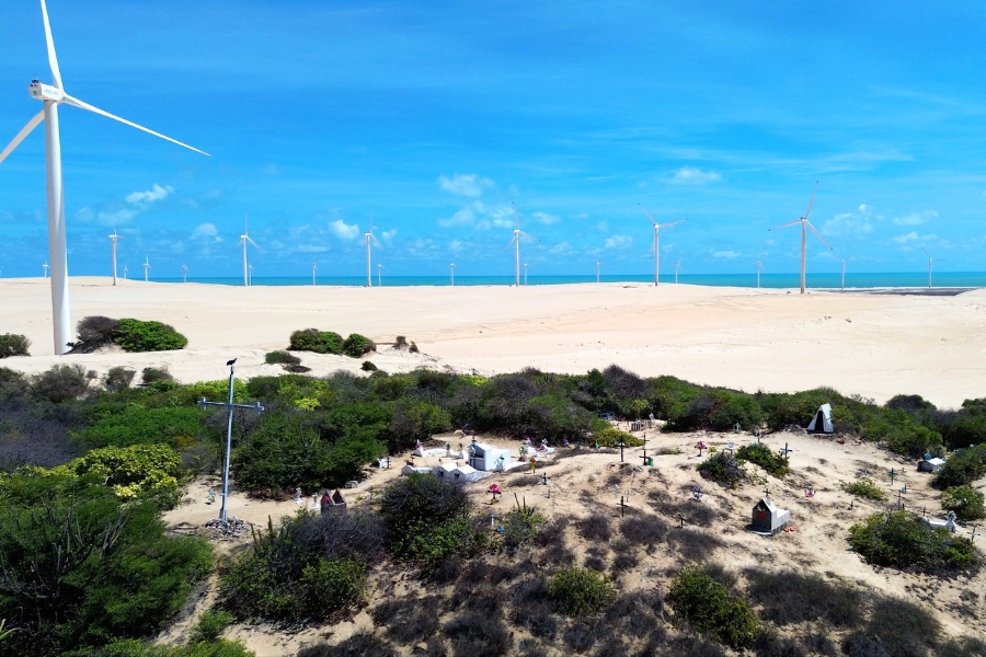Foto colorida de faixa de praia com área coberta de vegetação e vários túmulos formando um cemitério tendo próximo turbinas eólicas e ao fundo o mar