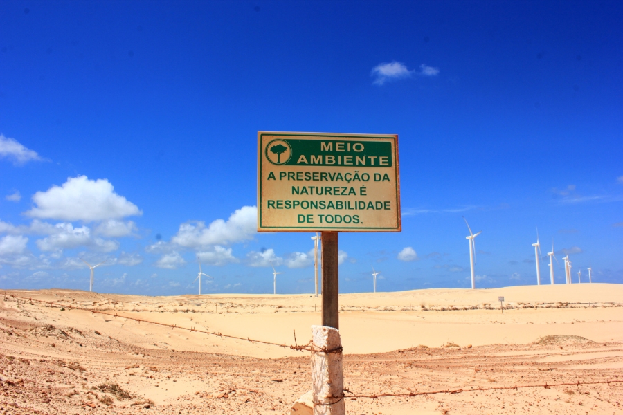 Foto colorida de praia com várias turbinas eólicas protegidas por cerca e com uma placa onde se lê meio ambiente a preservação da natureza é responsabilidade de todos