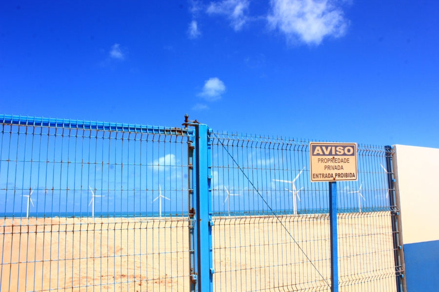 Foto colorida da beira do mar com uma cerca eletrificada isolando turbinas eólicas posicionadas na frente do mar. Na cerca se lê aviso propriedade privada entrada proibida