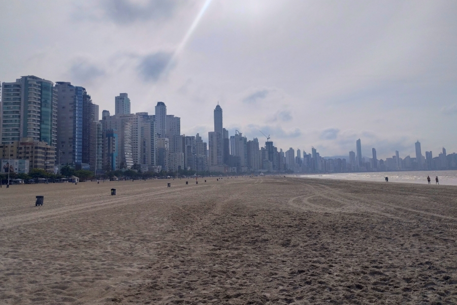 Foto de uma praia muito larga, com areia fofa que se estende até encontrar o mar calmo. À sua esquerda, uma fileira de prédios altíssimos forma uma densa parede urbana que acompanha toda a orla