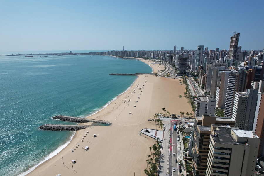 A foto mostra uma longa faixa de praia com areia clara e mar azul, com uma cidade ao lado, repleta de prédios altos ao longo da orla. Há quebra-mares de pedras entrando no mar e poucos guarda-sóis espalhados na areia