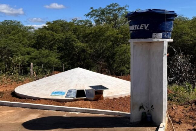 A imagem mostra uma cisterna de concreto em forma de cone achatado, parcialmente enterrada no chão, usada para armazenar água da chuva. Ao lado, há uma caixa d'água azul sobre uma coluna branca de concreto, cercada por vegetação do semiárido. na estrutura da cisterna foi instalada uma placa solar para geração de energia