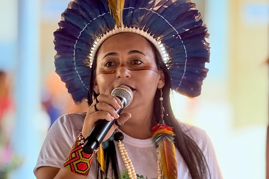 Uma mulher, possivelmente pertencente a um grupo indígena brasileiro, veste um cocar vibrante de penas azuis e amarelas e pinturas faciais tradicionais. Ela segura um microfone e parece estar falando ou cantando, com um colar de contas brancas e uma pulseira colorida adornando seu pulso