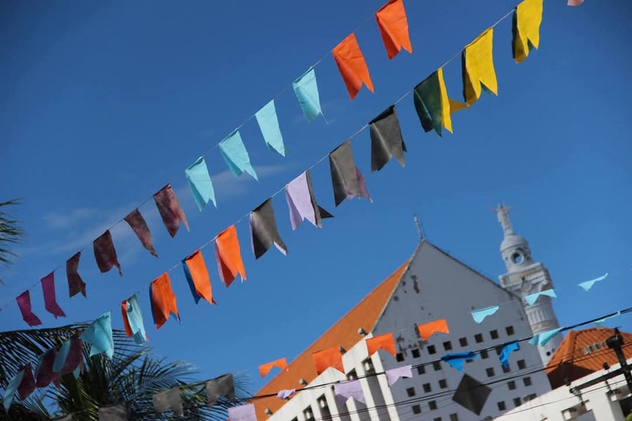 Bandeirolas coloridas enfeitam o céu azul em um dia ensolarado. Ao fundo, vê-se um prédio branco de telhado laranja e uma torre com cúpula, compondo um cenário típico de festa junina nordestina