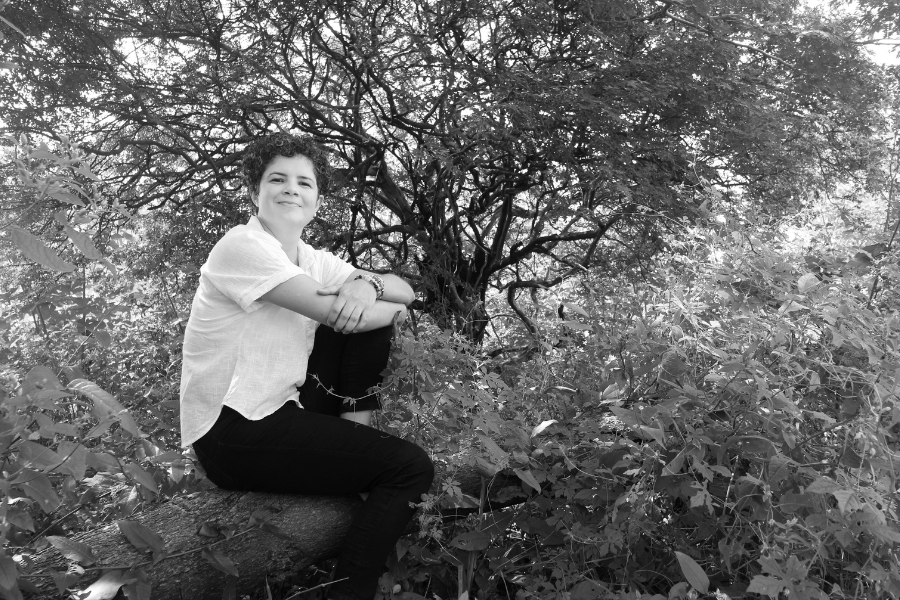 Foto em preto e branco de mulher branca de cabelos curtos e cacheados vestindo camisa de manga curta clara e calça escura está sentada sobre um tronco de árvore, cercada por uma vegetação densa. Ela sorri, com os braços apoiados em um dos joelhos, em um ambiente natural sombreado por galhos e folhas