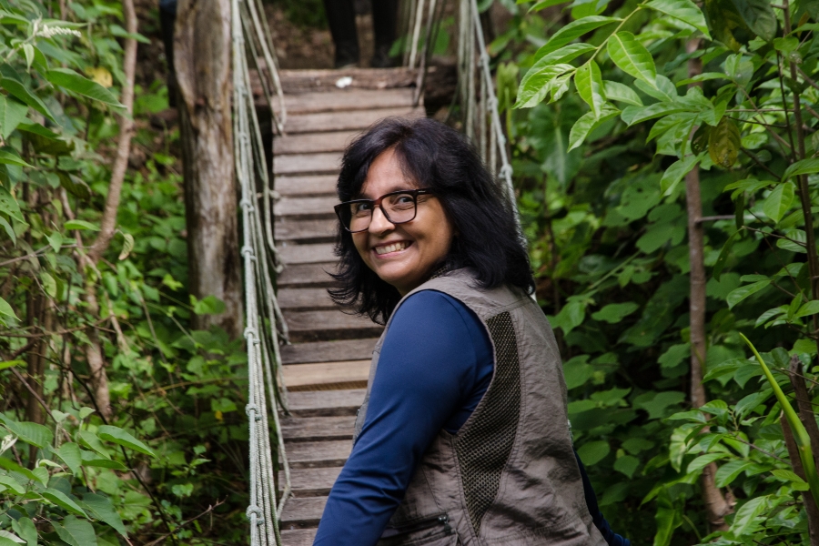 Uma mulher sorridente com óculos e colete bege olha para a câmera enquanto está em uma ponte suspensa de madeira em uma área verde exuberante. Outra pessoa pode ser vista subindo a ponte ao fundo