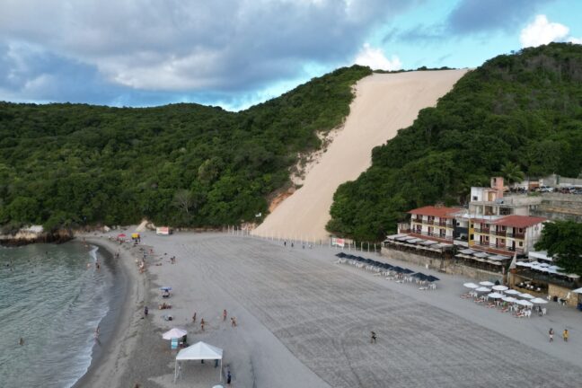 Foto de uma praia com faixa de areia larga e clara, banhada por águas calmas. À sua frente, uma enorme duna, de coloração e textura diferente, desce por uma encosta verde, cercada por vegetação densa e algumas construções à beira-mar