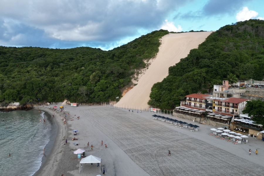 Foto de uma praia com faixa de areia larga e clara, banhada por águas calmas. À sua frente, uma enorme duna, de coloração e textura diferente, desce por uma encosta verde, cercada por vegetação densa e algumas construções à beira-mar