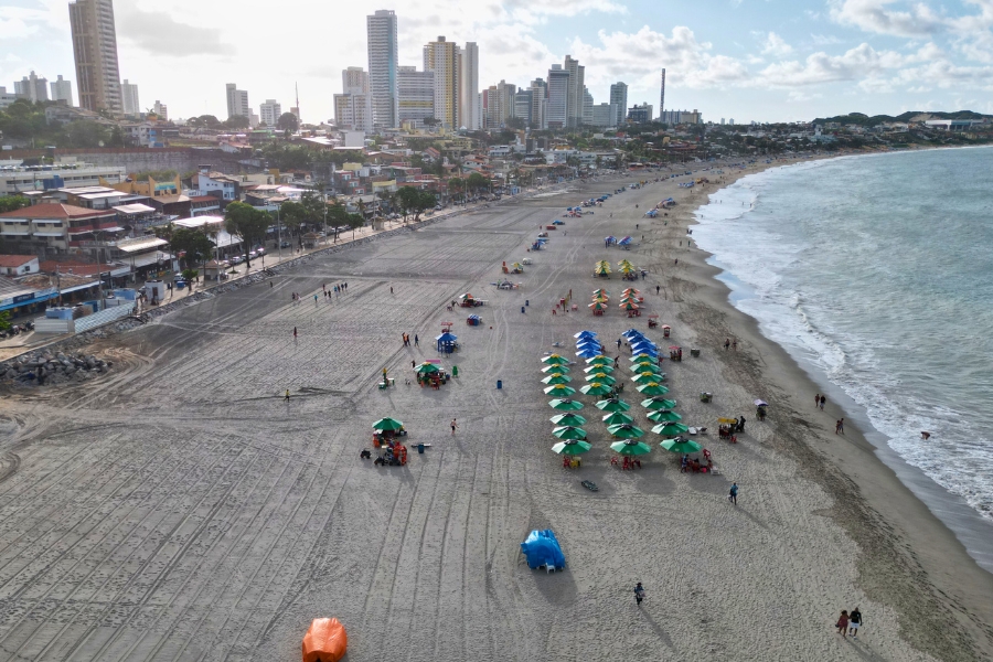 Foto de uma praia urbana ampla, com muitas fileiras de guarda-sóis coloridos e pessoas espalhadas pela areia. Ao fundo, há uma cidade com vários prédios altos próximos à orla