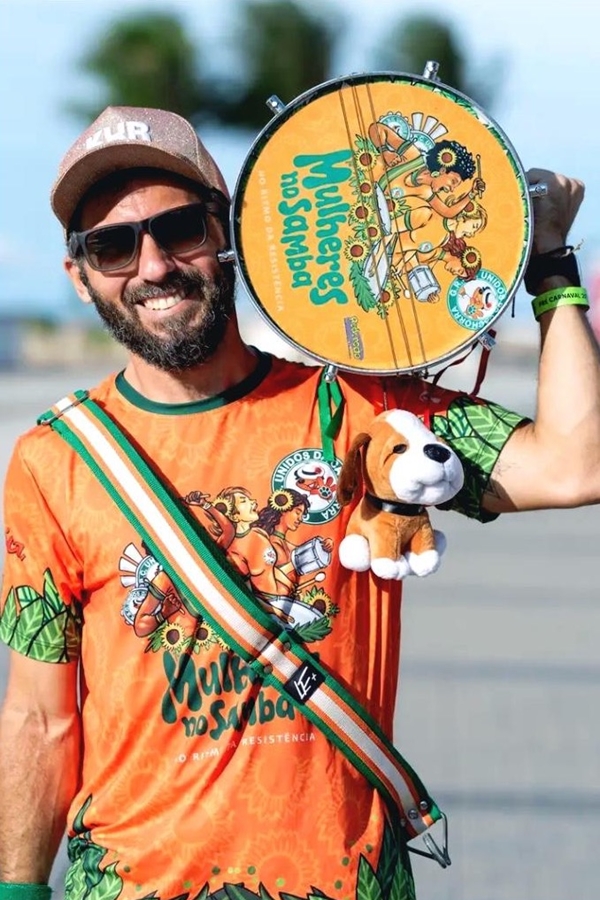 Homem com barbas, usando boné marrom, óculos escuros, um abadá de carnavall laranja. Ele segura um tambor