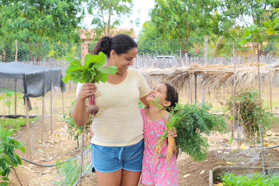 Uma mulher e uma menina sorriem olhando uma para a outra em uma horta. A mulher segura beterrabas frescas na mão direita e, com o braço esquerdo sobre os ombros da menina, segura, com a mão esquerda, cenouras com folhas, em meio a canteiros verdes e árvores ao fundo