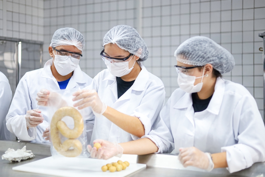 Três pessoas com jalecos brancos, toucas, máscaras, luvas e óculos de proteção trabalham em uma cozinha profissional. Duas modelam pequenas bolas de massa sobre uma bancada e uma segura um pacote plástico transparente com um biscoito em forma de oito. O ambiente tem paredes de azulejos brancos e iluminação forte